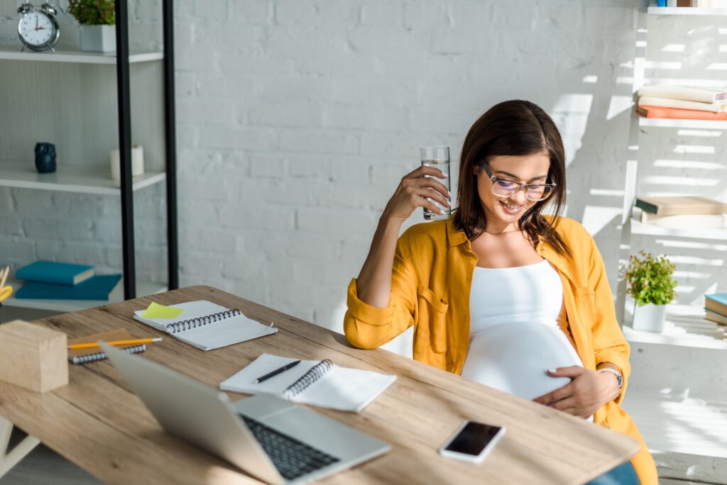 beautiful pregnant freelancer holding glass of water in office with home office